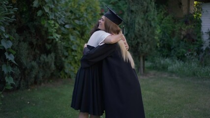Happy young graduate woman hugging with mature mother outdoors in garden. Smiling slim beautiful daughter embracing parent on graduation day on backyard. Education and family concept