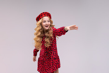 Little girl in french style hat points her finger to side. Happy girl with long curly hair in a red beret and dress. Parisian child on a light background. Summer fashion and beauty.