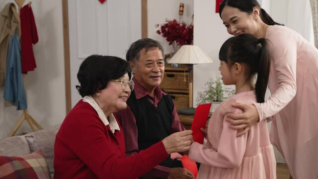 elderly couple grandmother and grandfather telling grandchild to come, giving red envelope lucky money and wishing her happy new year with hand gestures in living room. translation: congratulations