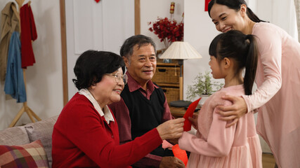 happy elderly couple grandmother and grandfather telling grandchild to come, giving red envelope lucky money and wishing her happy chinese new year with hand gestures in the living room at home