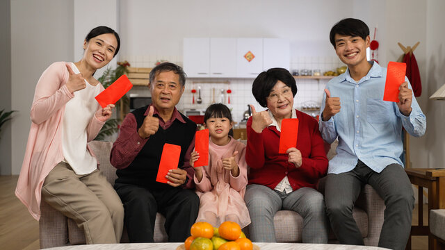 happy family of mixed generation smiling at camera with thumb and victory hand gestures while taking burst photos with red envelopes lucky money at home on Chinese new year
