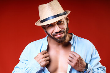 Cuban or Latino with tanned body in hat and shirt poses on red background. Studio portrait of bearded man 30-40 years old. Brutal man looks into camera and smiles. Summer vacation.