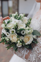 Caucasian bride in a veil holds a wedding bouquet close to her chest, a close-up photo against the background of a white wedding dress decorated with white shiny embroidery, close-up photo