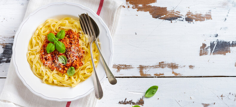 Traditional Italian dish fettuccine pasta with bolognese sauce, basil and parmesan cheese in a white plate on a light wooden background. Top view.