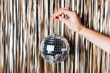 Woman hand is holding disco ball in the middle of Golden foil tinsel strips. Festive background for christmas, new year, holidays or birthday celebration. Creative art concept.
