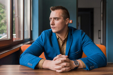 A brooding, young man of European appearance portrait businessman in jacket and shirt sits idly by at a table in a cafe and looks out the window