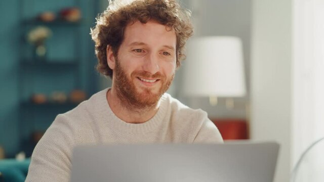 Handsome Adult Man Portrait with Ginger Curly Hair Using Laptop Computer, Sitting in Living Room in Apartment. Happy Man is Working from Home, Online Shopping, Watching Videos or Writing Emails.