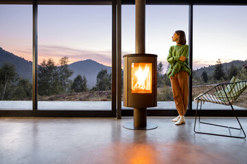 Woman enjoy great view on mountains while sitting near fireplace at modern living room at sunset ....