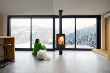 Woman sitting with dog near fireplace and panoramic window at modern living room with stunning view...