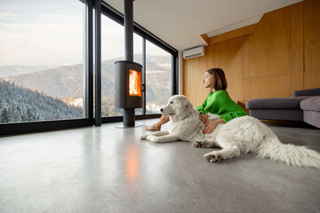 Woman sitting with dog near fireplace and panoramic window at modern living room with stunning view on snowy mountains. Concept of rest in houses or cabins on nature. Idea of escape from everyday life