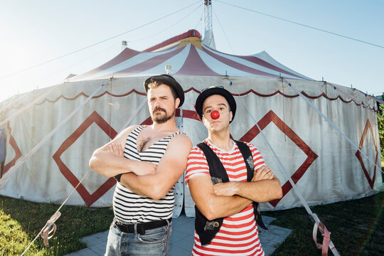 Muscular build artist standing with arms crossed by clown in front of circus tent