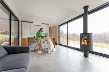 Young woman with her dog at modern house with panoramic windows and fireplace. Concept of comfort and happy life at modern living spaces. Wide interior view