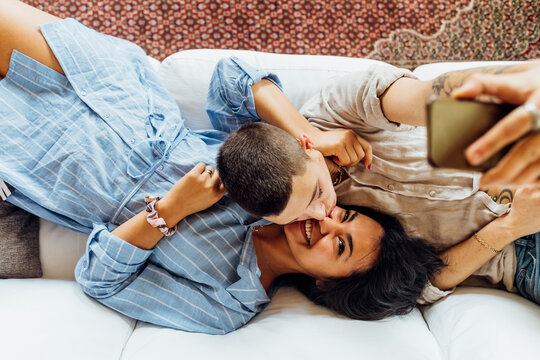 Affectionate woman kissing girlfriend while taking selfie through smart phone at home