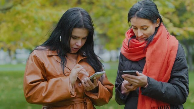 Two young women holding telephones in hands closeup looking at gadgets one helps other download cool free mobile app hispanics girls stand in autumn park weekend girlfriends discussing online shopping