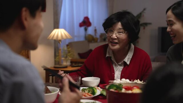 selective focus of a cheerful asian senior woman grandmother wearing red clothing talking with hand gestures while chatting with her adult son at dinner table during family reunion meal at home