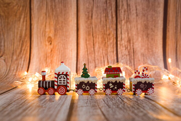 A festive train carries gifts for Christmas. A red toy train. Wooden background with Christmas lights.  Space for text. Christmas card.