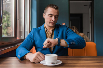 A busy business man a stylish businessman of Caucasian appearance in a suit and shirt watches the time and is late for negotiations, sitting at a cup of coffee at a table in a cafe