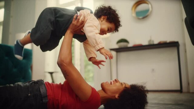 Beautiful Black Latina Mother Playing with Adorable Baby Boy at Home on Living Room Floor. Cheerful Mom Nurturing a Child. Happy Son Raised in the Air. Concept of Childhood, New Life, Parenthood.