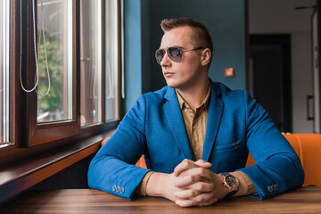 A young businessman of European appearance stylish portrait with sunglasses sits at a table in a cafe idly by at look at the window