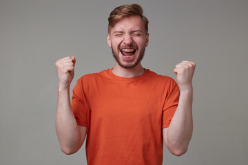 Studio portrait of young bearded man, wears casually posing over gray studio background celebrate a goal of his favorite team. shows victory sign. isolated.