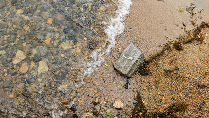 a rectangular stone is tied with a white rope, like a gift. lies on a rocky shore next to the water. waves wash over him. a gift for the summer