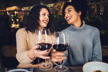 Multiracial couple of young best friends celebrating toasting with red wine - multicultural girlfriends clinking wineglasses in restaurant