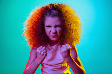 Close-up little red-headed child, disgusted girl looking at camera isolated on blue studio background in purple neon light.