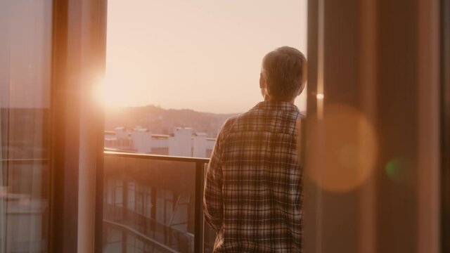 Casual bearded adult man spending morning home, enjoying beautiful senset from balcony and drinking coffee. Cozy apartment. Sceneries. Urban landscape.
