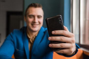 Close-up, businessman guy holds a black smartphone or mobile phone in his hand and takes a selfie or talks on a video link in a cafe