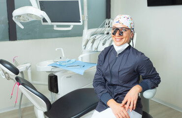 Beautiful female dentist sitting near the dental chair. Portrait with personal protective equipment and binocular glasses.