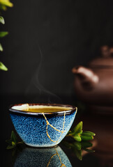 Bowl of green Japanese tea, tea leaves lie next the cup. Selective focus on the cup. Steam rises above the bowl. Reclaimed ceramic blue cup, second life of things, recycling or kintsugi