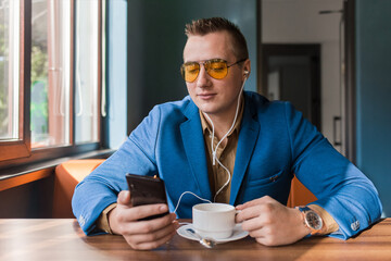 A businessman a stylish portrait of Caucasian appearance in sunglasses, suit style, sits at a table on a coffee break in smartphone and headphones, listen to music on cellphone or mobile in a cafe