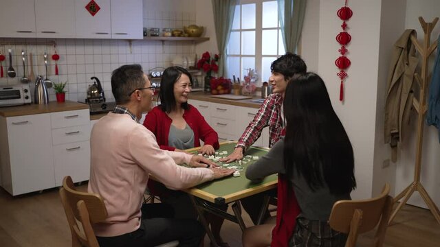smiling asian family of four having fun with mahjong game and chatting lively in the evening at home during Chinese new year holiday. chinese text at background translation: luck