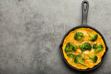 Healthy vegetarian egg omelette with green broccoli in black cast iron frying pan with fork and knife top view on gray stone concrete background table, diet and healthy clean eating concept copy space