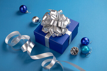 Christmas present box wrapped in blue paper with silver ribbon and bow, Christmas sparkly round balls decorations on blue paper background angle view, Christmas composition in blue and silver colors