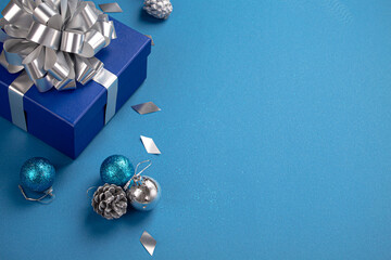 Christmas present box wrapped in blue paper with silver ribbon and bow, Christmas round balls decorations on blue paper background with copy space, Christmas composition in blue and silver colors