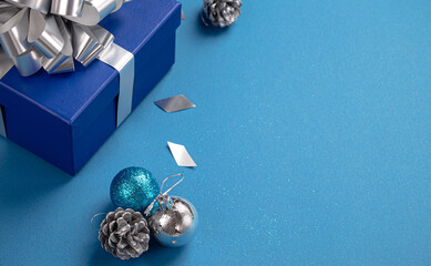 Christmas present box wrapped in blue paper with silver ribbon and bow, Christmas round balls decorations on blue paper background with copy space, Christmas composition in blue and silver colors