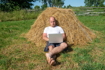 Naklejka na ściany i meble a middle-aged man, dressed in a T-shirt and shorts, is lying on a yellow haystack with a gray laptop in his hands. Remote work, training. Rural area. Thoughtful facial expression, front view