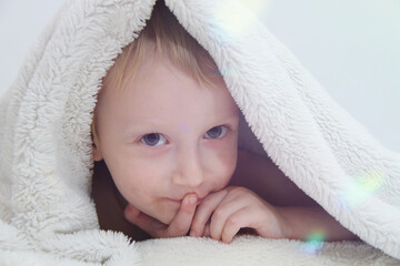 Cozy holidays at home. Cute little child lying under white knitted blanket and playing