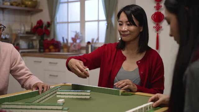 selective focus of smiling mother drawing keeping a tile and discarding another from her hand while playing mahjong game at home with family on Chinese new year's eve