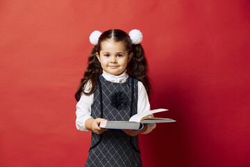 Portrait of a little girl in school uniform looking at camera and holding an open book. - obrazy, fototapety, plakaty