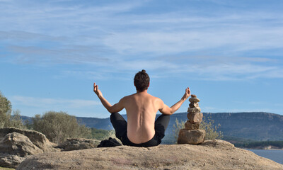 Man from behind with naked torso doing yoga, fingers in mudra position. Stack of stones next to the...