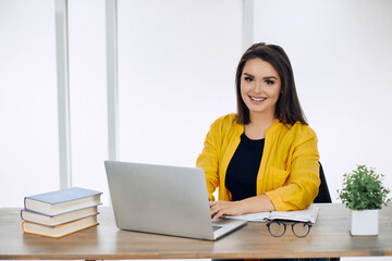 Young smiling attractive office worker woman sitting at her work place. A table with a laptop some books glasses plant and a bright window behind.