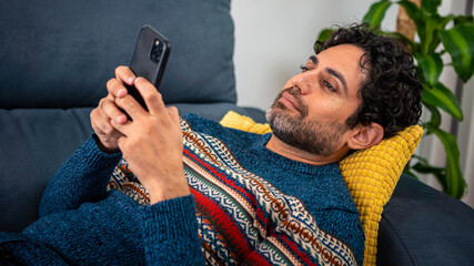 Cheerful handsome man using his smart phone while lying down on couch at home
