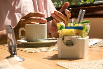 Man's hand holding a white ceramic cup or mug with hot beverage like coffee or tea. Sugar and adulcorants unfocused on foreground. Wooden table, mand with pink shirt