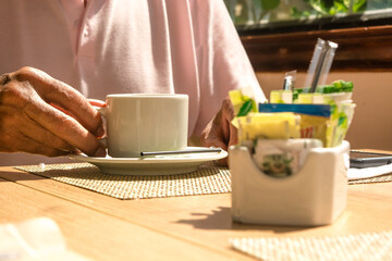 Man's hand holding a white ceramic cup or mug with hot beverage like coffee or tea. Sugar and adulcorants unfocused on foreground. Wooden table, mand with pink shirt