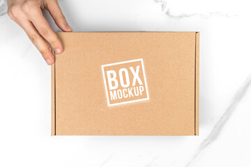 Cardboard box mockup in delivery man hand, top view. White table background. Suitable for food, cosmetic or medical packaging. Shipping and delivery concept