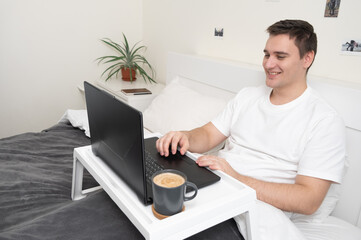 Handsome smiling man working on laptop in bed. Freelancer at home. A programmer or developer works remotely.