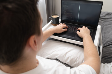 Handsome smiling man working on laptop in bed. Freelancer at home. A programmer or developer works remotely.