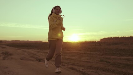 girl in headphones runs at sunset sky, listening to music for active training, speakers in ears of triathlon athlete, happy female morning jog for cardio movement, healthy body life, fitness exercise